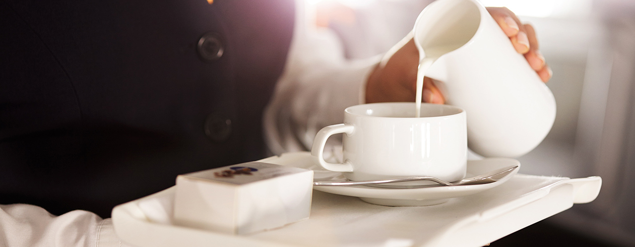 Nahaufnahme eines Tabletts in den Händen einer Flugbegleiter:in, welche Milch in eine Tasse Kaffee gießt