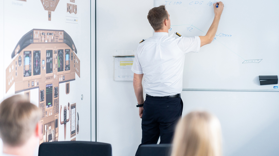 In a seminar room a pilot as instructor writes something on a whiteboard while two people watch