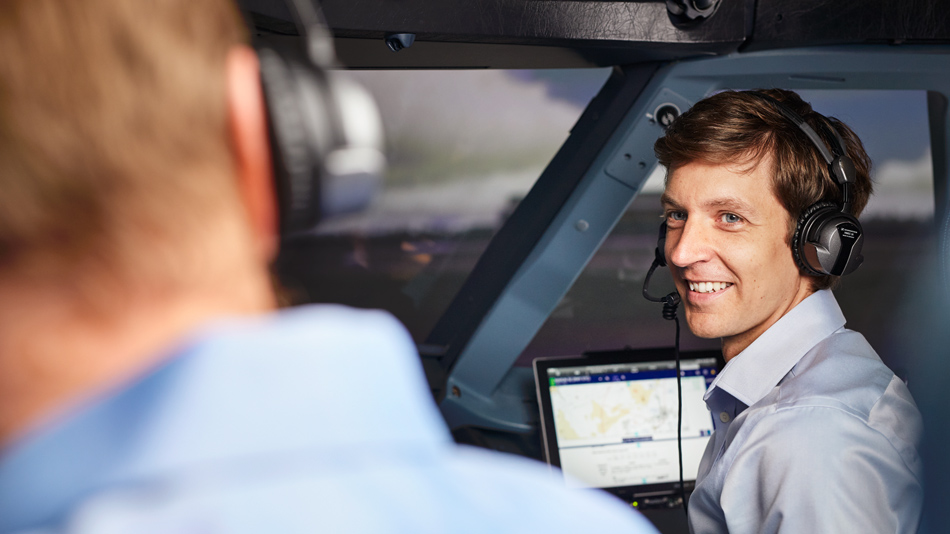 Ein Mann, der Kopfhörer aufhat, sitzt in einem Flugzeugcockpit und schaut lächelnd zu seinem Kollegen