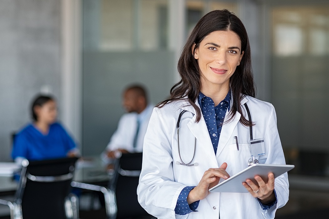 Eine Ärztin steht mit Tablet und Stethoskop im Vordergrund, im Hintergrund sitzen zwei Personen.